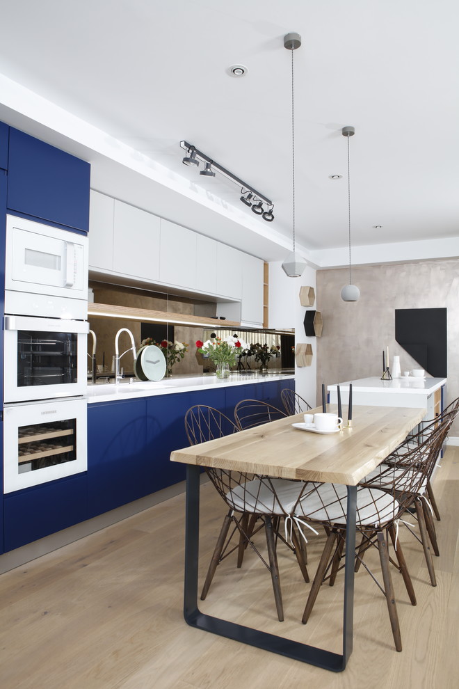 Zweizeilige Moderne Wohnküche mit flächenbündigen Schrankfronten, blauen Schränken, Rückwand aus Spiegelfliesen, weißen Elektrogeräten, hellem Holzboden, Kücheninsel, beigem Boden und weißer Arbeitsplatte in Sonstige