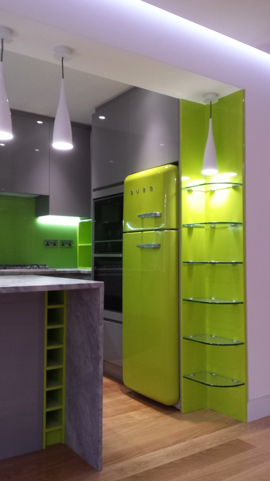 Small contemporary u-shaped kitchen/diner in London with flat-panel cabinets, grey cabinets, granite worktops, green splashback, glass sheet splashback, light hardwood flooring, a breakfast bar, a built-in sink and stainless steel appliances.