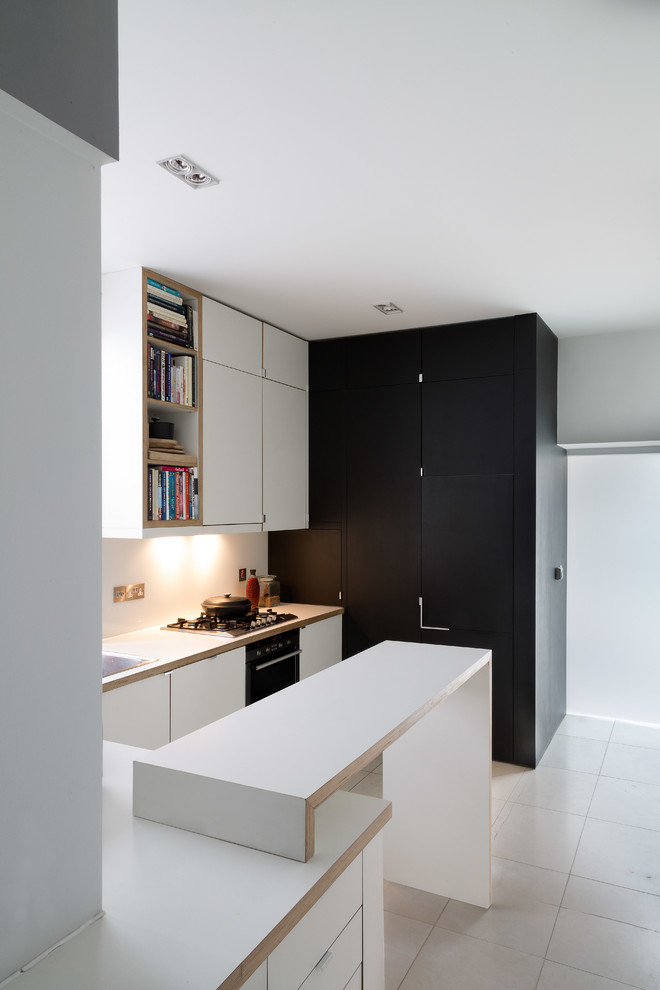 Large contemporary l-shaped open plan kitchen in London with a built-in sink, flat-panel cabinets, white cabinets, laminate countertops, white splashback, glass sheet splashback, black appliances, limestone flooring and a breakfast bar.