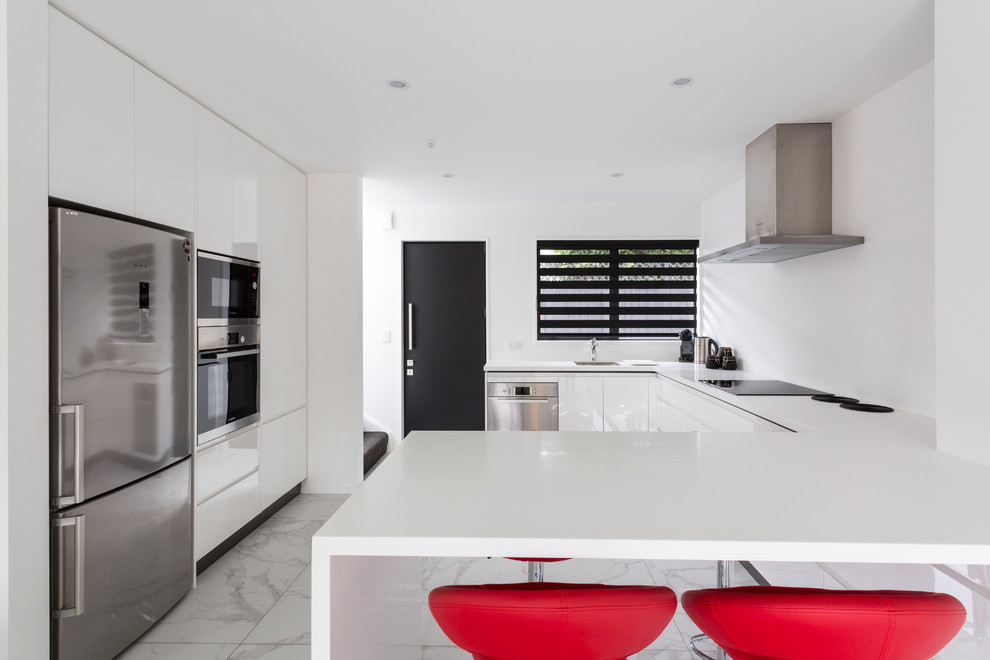Minimalist u-shaped porcelain tile and multicolored floor open concept kitchen photo in Christchurch with a single-bowl sink, flat-panel cabinets, white cabinets, solid surface countertops, white backsplash, glass sheet backsplash, stainless steel appliances, a peninsula and white countertops