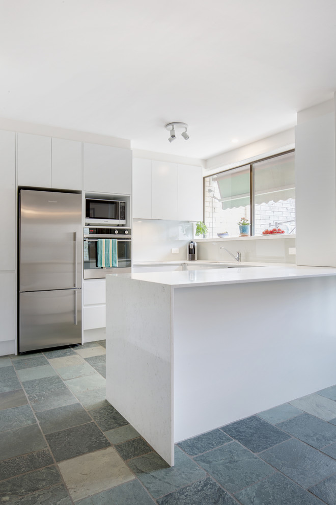 Mid-sized minimalist u-shaped slate floor and blue floor eat-in kitchen photo in Sydney with an undermount sink, flat-panel cabinets, white cabinets, quartz countertops, white backsplash, glass sheet backsplash, stainless steel appliances, a peninsula and white countertops