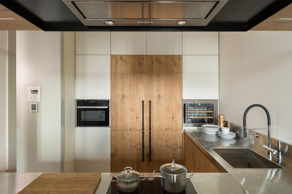 Foto på ett funkis grå kök, med släta luckor, skåp i mellenmörkt trä, bänkskiva i rostfritt stål, grått stänkskydd, rostfria vitvaror, en halv köksö och en enkel diskho