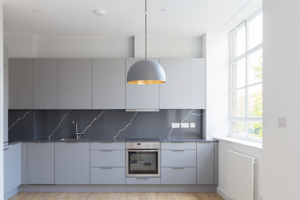 Medium sized contemporary l-shaped open plan kitchen in London with a built-in sink, flat-panel cabinets, grey cabinets, composite countertops, grey splashback, marble splashback, stainless steel appliances, light hardwood flooring, no island, brown floors and grey worktops.