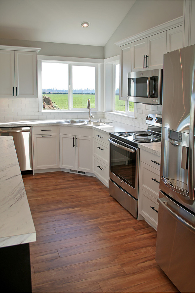 Example of a large arts and crafts l-shaped medium tone wood floor open concept kitchen design in Seattle with a drop-in sink, recessed-panel cabinets, white cabinets, laminate countertops, white backsplash, ceramic backsplash, stainless steel appliances and an island