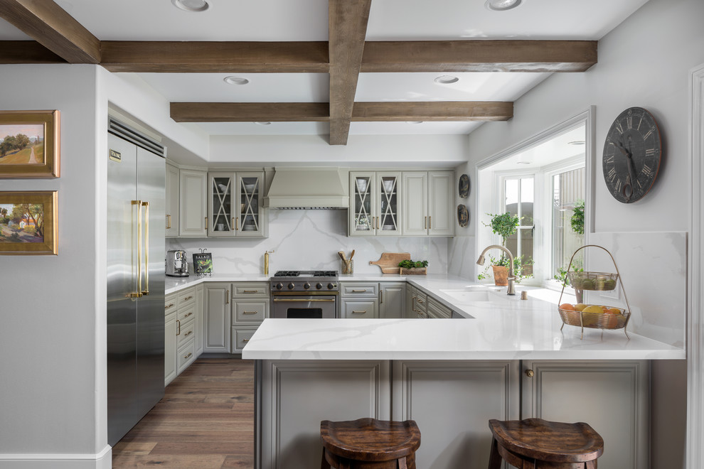 Inspiration for a traditional u-shaped kitchen in Orange County with a submerged sink, raised-panel cabinets, grey cabinets, white splashback, stone slab splashback, stainless steel appliances, medium hardwood flooring, a breakfast bar, brown floors and white worktops.