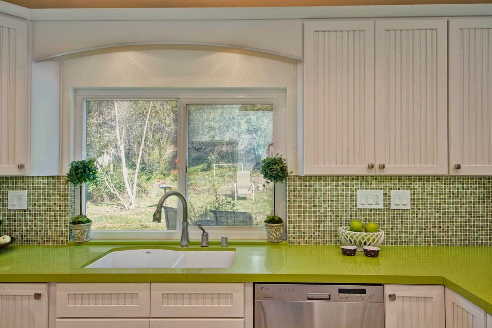 Cette photo montre une cuisine américaine chic en U avec un évier encastré, un placard à porte affleurante, des portes de placard blanches, un plan de travail en quartz modifié, une crédence verte et un électroménager en acier inoxydable.