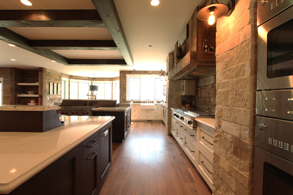 This is an example of a large farmhouse l-shaped open plan kitchen in Other with a belfast sink, recessed-panel cabinets, dark wood cabinets, engineered stone countertops, multi-coloured splashback, stone slab splashback, integrated appliances, medium hardwood flooring, multiple islands, brown floors and white worktops.