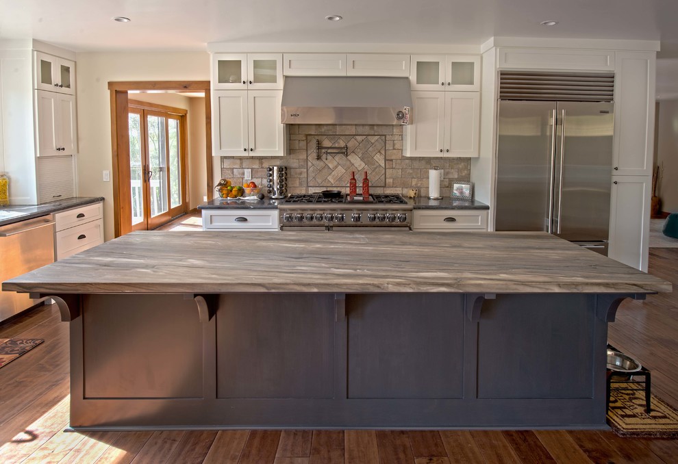 Large arts and crafts l-shaped dark wood floor and brown floor open concept kitchen photo in San Francisco with a farmhouse sink, shaker cabinets, white cabinets, granite countertops, beige backsplash, stone tile backsplash, stainless steel appliances, an island and multicolored countertops