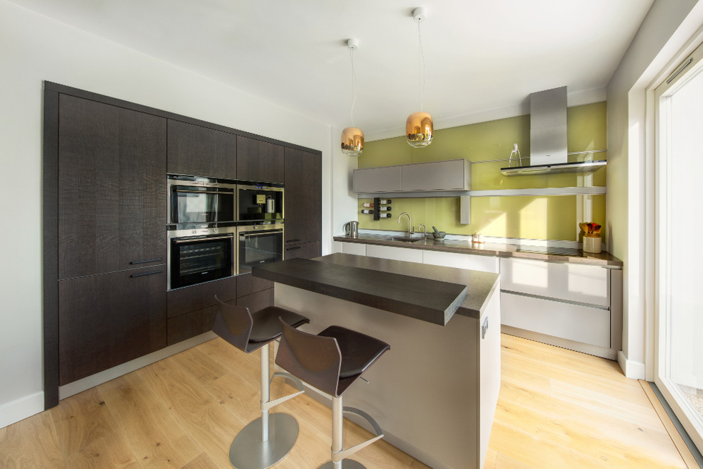 Kleine Moderne Wohnküche in L-Form mit integriertem Waschbecken, flächenbündigen Schrankfronten, grauen Schränken, Mineralwerkstoff-Arbeitsplatte, Küchenrückwand in Grün, Glasrückwand, schwarzen Elektrogeräten, hellem Holzboden, Kücheninsel und grauer Arbeitsplatte in Oxfordshire