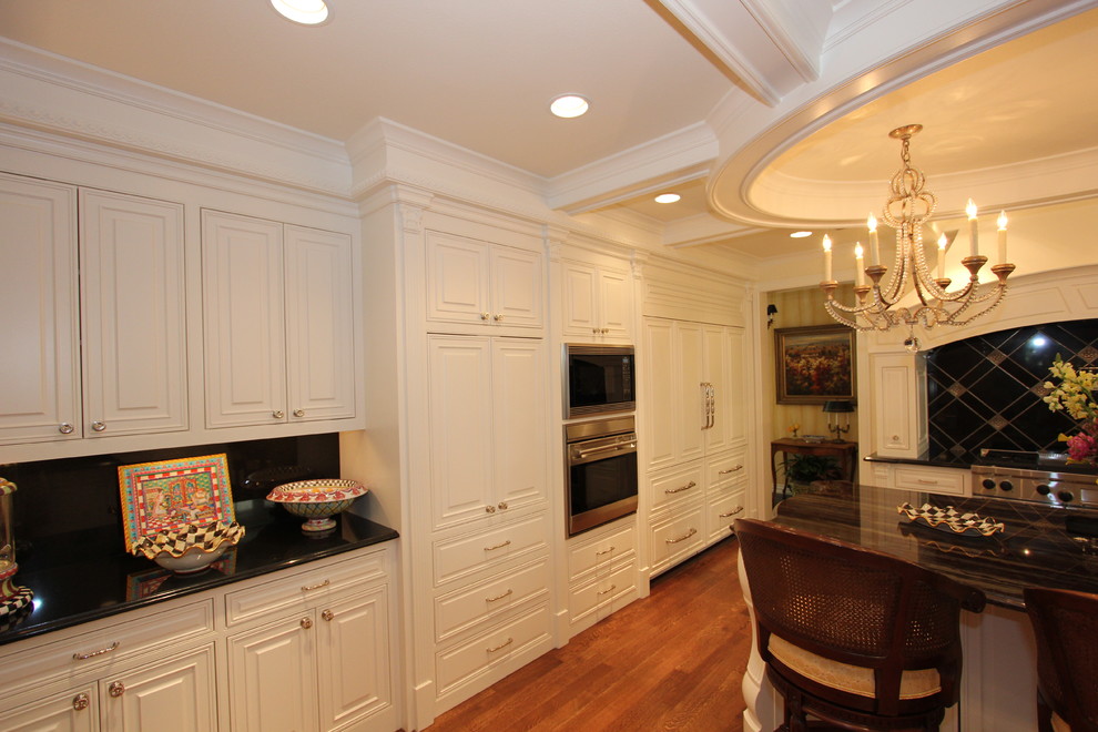 Exempel på ett mellanstort klassiskt kök, med en dubbel diskho, luckor med upphöjd panel, vita skåp, granitbänkskiva, svart stänkskydd, stänkskydd i sten, rostfria vitvaror, mellanmörkt trägolv, en köksö och brunt golv