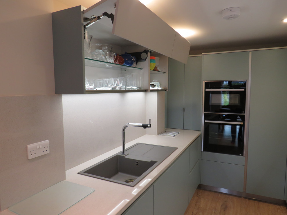 Inspiration for a small contemporary u-shaped enclosed kitchen in Essex with a built-in sink, flat-panel cabinets, green cabinets, quartz worktops, beige splashback, black appliances, light hardwood flooring, no island, brown floors and beige worktops.