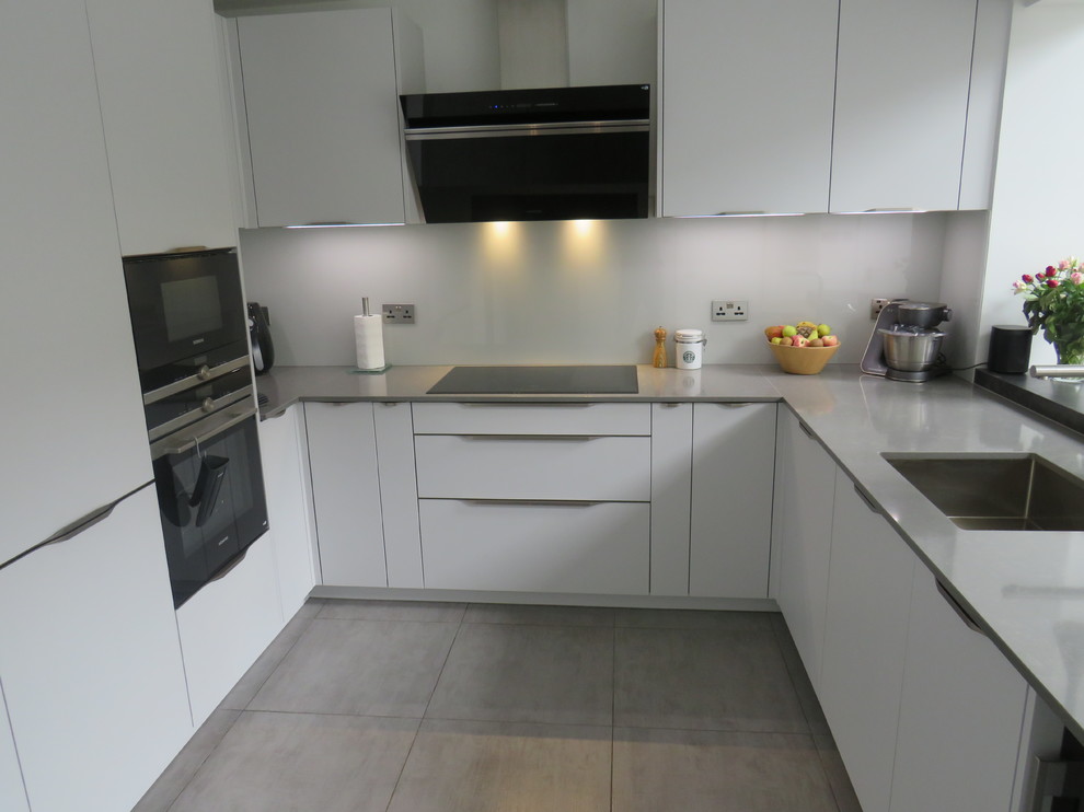 This is an example of a medium sized contemporary u-shaped kitchen/diner in Essex with a submerged sink, flat-panel cabinets, grey cabinets, quartz worktops, grey splashback, glass sheet splashback, black appliances, porcelain flooring, a breakfast bar, grey floors and grey worktops.