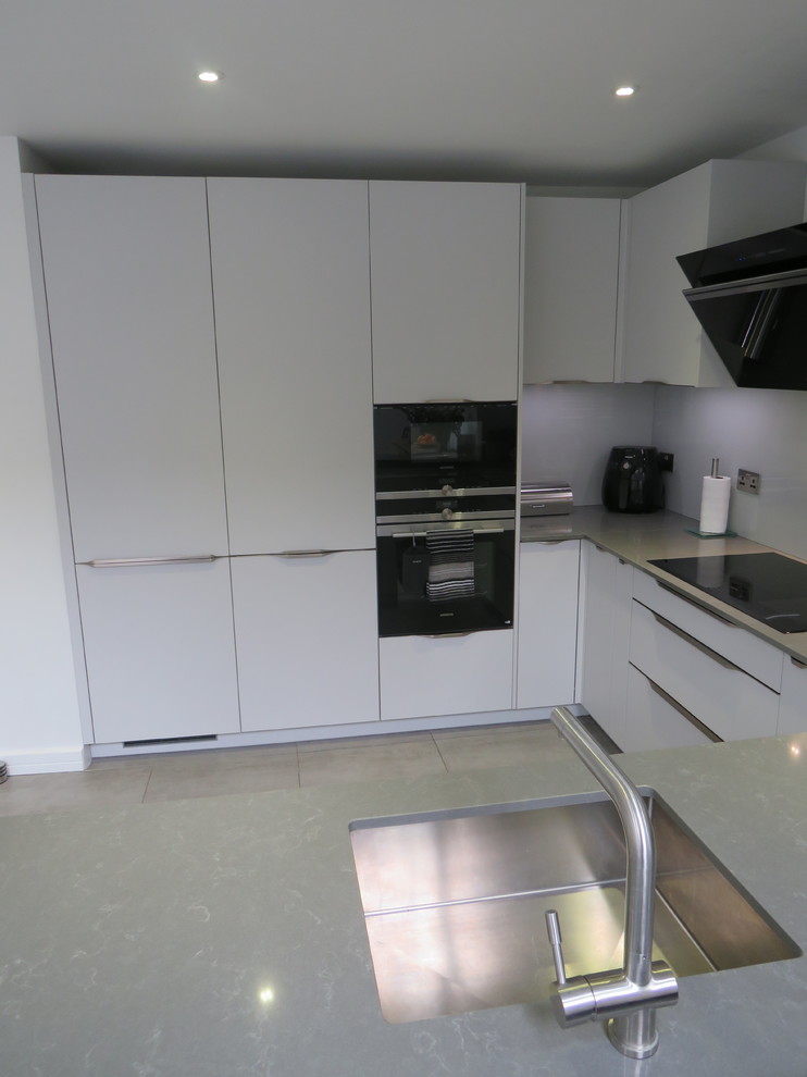 Bild på ett mellanstort funkis grå grått kök, med en undermonterad diskho, släta luckor, grå skåp, bänkskiva i kvartsit, grått stänkskydd, glaspanel som stänkskydd, svarta vitvaror, klinkergolv i porslin, en halv köksö och grått golv