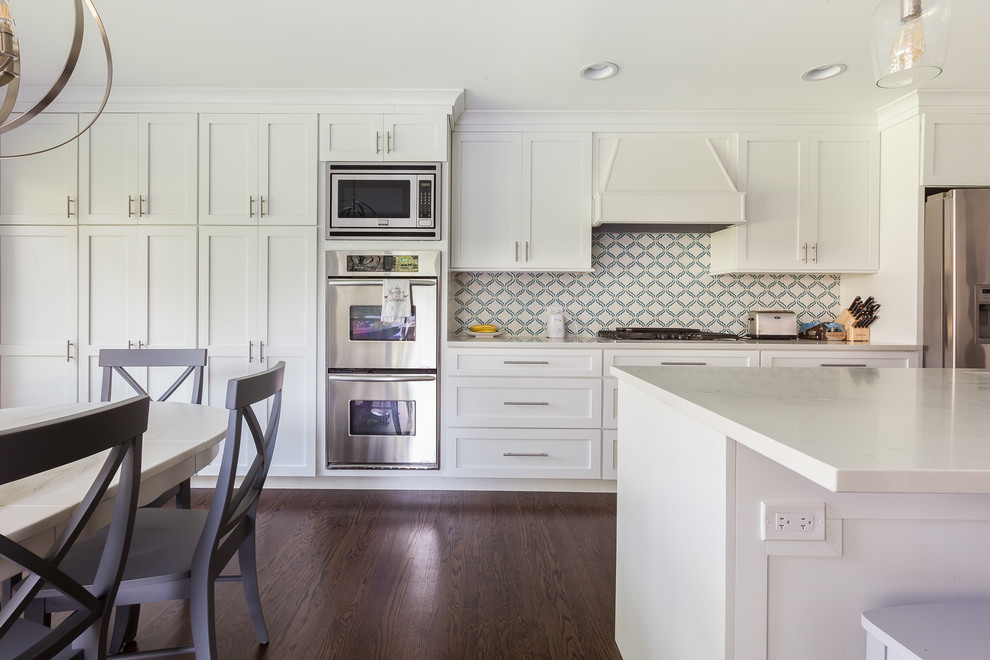 Bild på ett stort funkis vit vitt kök, med en undermonterad diskho, skåp i shakerstil, vita skåp, bänkskiva i kvarts, vitt stänkskydd, rostfria vitvaror, mellanmörkt trägolv, en köksö och brunt golv