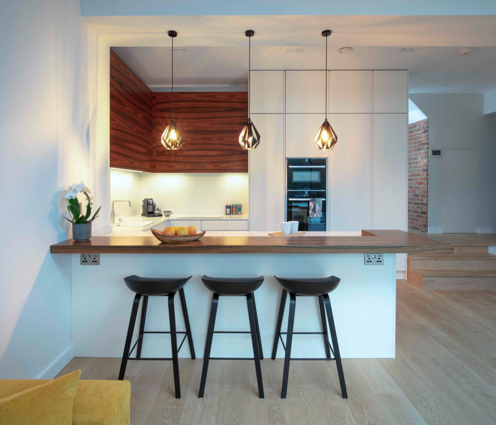 Design ideas for a small contemporary l-shaped kitchen/diner in London with an integrated sink, white splashback, black appliances, light hardwood flooring, an island, beige floors, white worktops, flat-panel cabinets and brown cabinets.