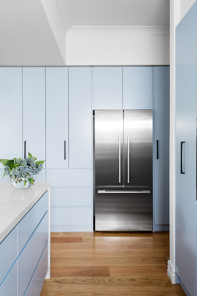 Inredning av ett modernt kök, med en nedsänkt diskho, släta luckor, blå skåp, stänkskydd med metallisk yta, spegel som stänkskydd, rostfria vitvaror, ljust trägolv och en halv köksö