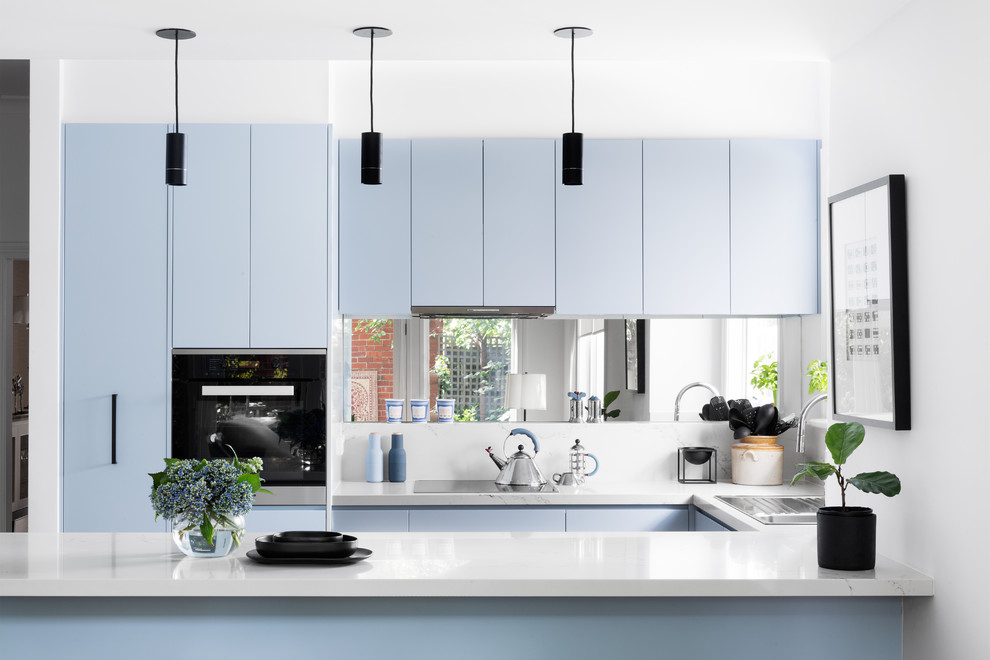 This is an example of a contemporary u-shaped open plan kitchen in Melbourne with a built-in sink, flat-panel cabinets, blue cabinets, metallic splashback, mirror splashback, stainless steel appliances, light hardwood flooring and a breakfast bar.