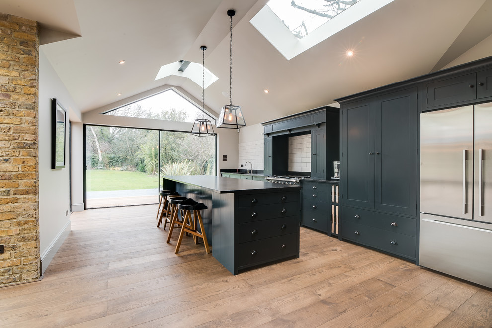 Inspiration for a large country galley kitchen/diner in Surrey with a built-in sink, shaker cabinets, blue cabinets, white splashback, ceramic splashback, stainless steel appliances, medium hardwood flooring, an island, brown floors and black worktops.