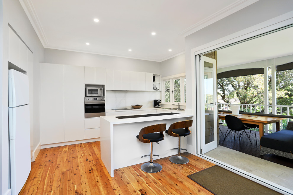 Design ideas for a medium sized contemporary u-shaped open plan kitchen in Sydney with a built-in sink, flat-panel cabinets, white cabinets, engineered stone countertops, white splashback, stone slab splashback, stainless steel appliances, medium hardwood flooring, a breakfast bar, yellow floors and white worktops.