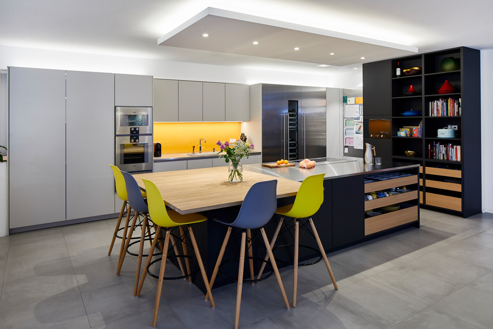 Inspiration for a large modern single-wall open plan kitchen with yellow splashback, an island, stainless steel appliances, grey floors, black cabinets, stainless steel worktops, glass sheet splashback and ceramic flooring.