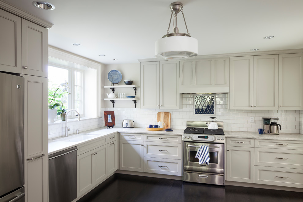 Foto på ett amerikanskt vit l-kök, med vita skåp, bänkskiva i kvarts, vitt stänkskydd, stänkskydd i tunnelbanekakel, rostfria vitvaror och mörkt trägolv