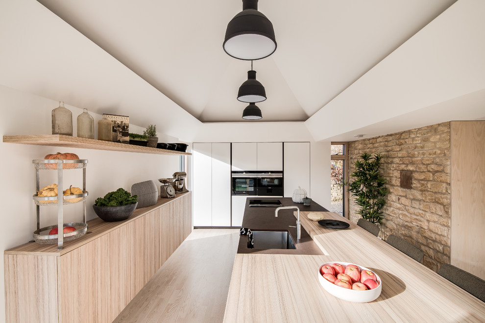 Bild på ett mellanstort funkis brun linjärt brunt kök med öppen planlösning, med en integrerad diskho, släta luckor, vita skåp, bänkskiva i koppar, svarta vitvaror, ljust trägolv, en halv köksö och beiget golv