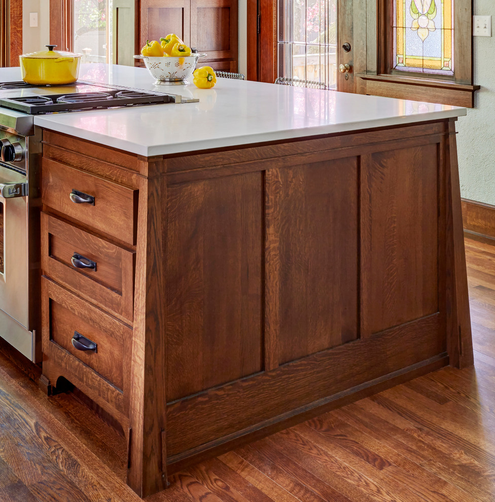 This is an example of a medium sized traditional kitchen in Milwaukee with a belfast sink, shaker cabinets, medium wood cabinets, engineered stone countertops, white splashback, metro tiled splashback, stainless steel appliances, medium hardwood flooring, an island, brown floors and white worktops.