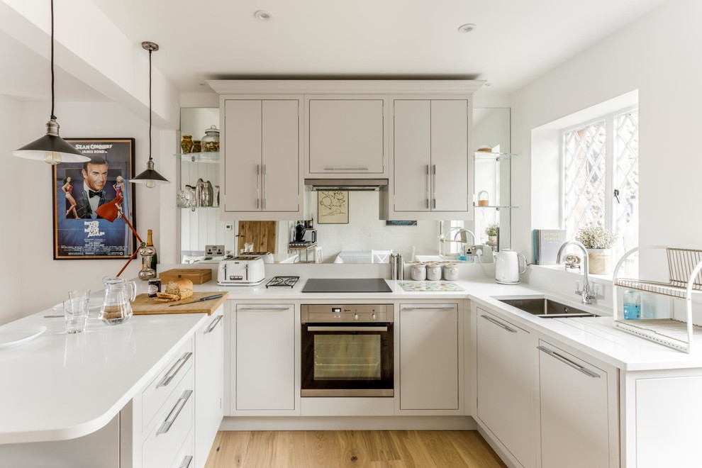 Inspiration for a classic u-shaped kitchen in Sussex with a double-bowl sink, flat-panel cabinets, beige cabinets, mirror splashback, light hardwood flooring, a breakfast bar, beige floors and white worktops.
