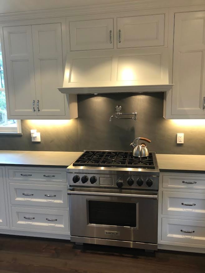 Large beach style l-shaped enclosed kitchen in New York with a belfast sink, recessed-panel cabinets, white cabinets, concrete worktops, grey splashback, stone slab splashback, stainless steel appliances, dark hardwood flooring, a breakfast bar, brown floors and grey worktops.