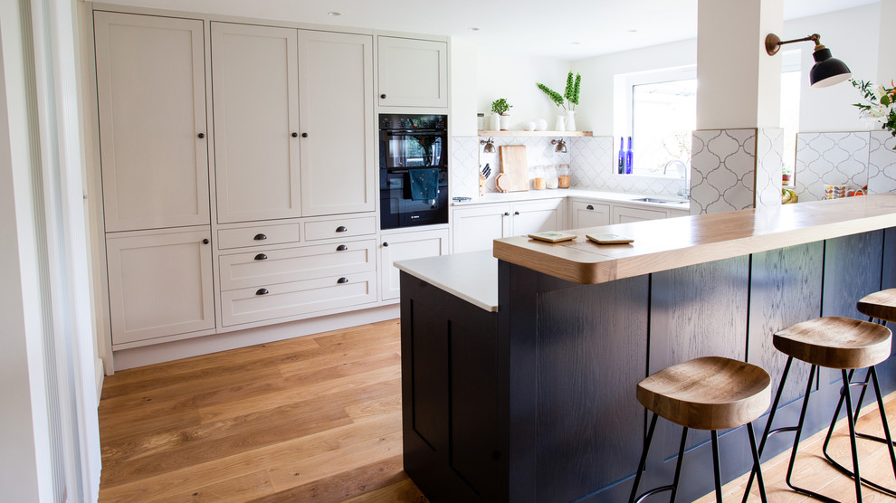 This is an example of a medium sized contemporary u-shaped kitchen in Other with a double-bowl sink, shaker cabinets, beige cabinets, white splashback, ceramic splashback, stainless steel appliances, light hardwood flooring, a breakfast bar, brown floors and white worktops.