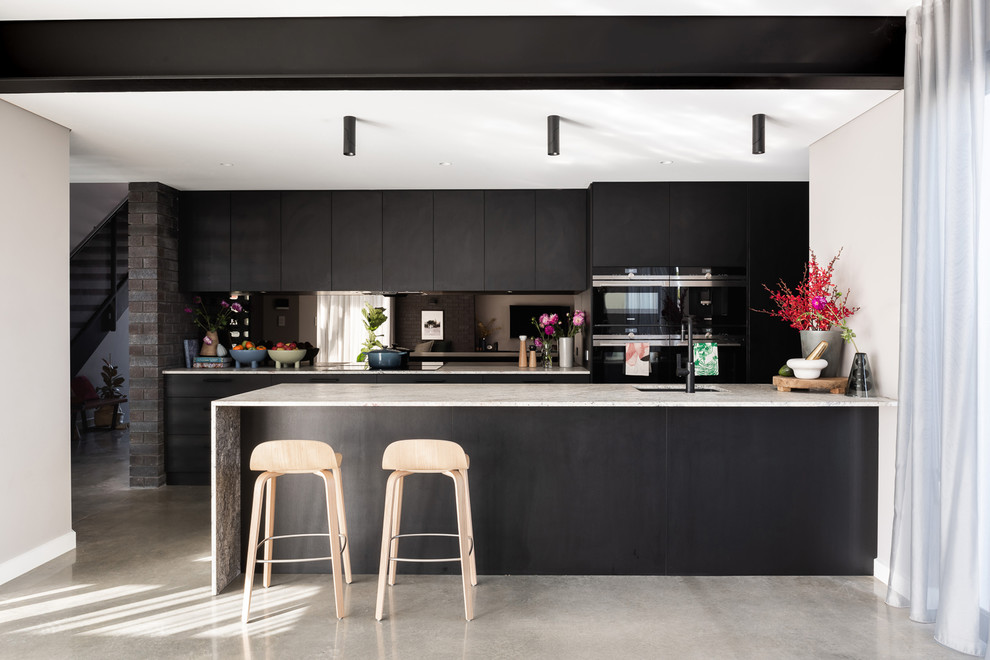 Bild på ett mellanstort funkis grå grått kök, med en enkel diskho, svarta skåp, granitbänkskiva, stänkskydd med metallisk yta, spegel som stänkskydd, svarta vitvaror, betonggolv, en köksö och grått golv