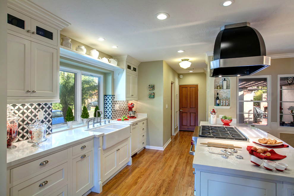 Photo of a medium sized traditional kitchen/diner in San Diego with a belfast sink, shaker cabinets, white cabinets, engineered stone countertops, multi-coloured splashback, integrated appliances, light hardwood flooring and a breakfast bar.