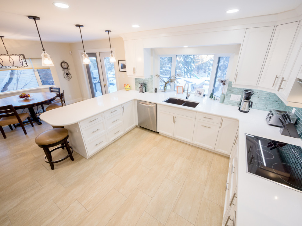 This is an example of a modern u-shaped kitchen/diner in Edmonton with a double-bowl sink, shaker cabinets, white cabinets, blue splashback, glass tiled splashback, porcelain flooring, quartz worktops, stainless steel appliances, a breakfast bar, beige floors and white worktops.