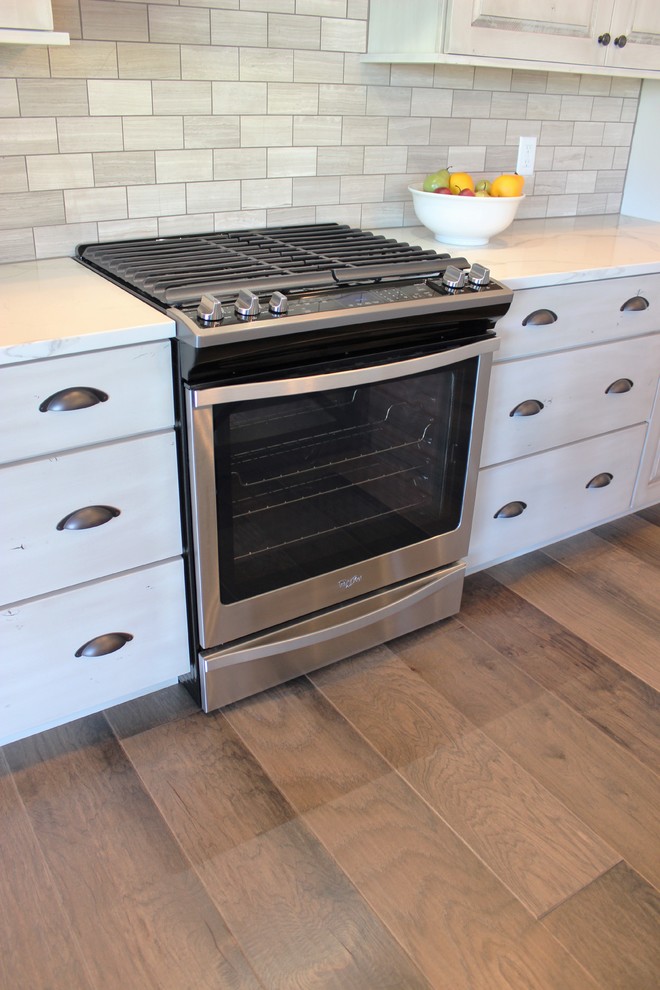 Bild på ett lantligt kök, med en rustik diskho, luckor med upphöjd panel, skåp i slitet trä, bänkskiva i kvarts, grått stänkskydd, stänkskydd i stenkakel, rostfria vitvaror, mellanmörkt trägolv, en köksö och grått golv
