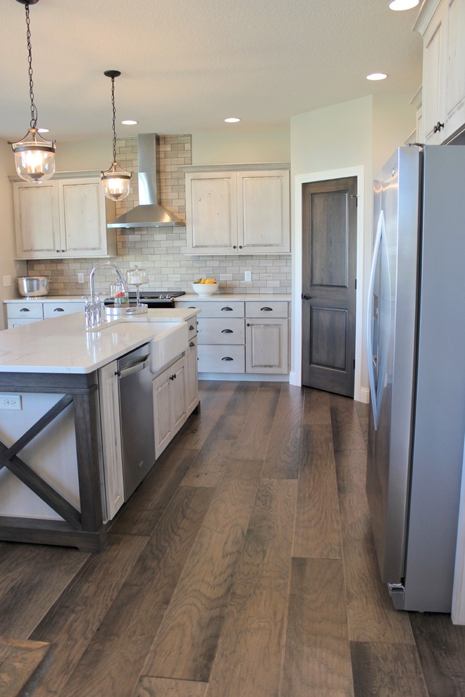 Example of a farmhouse l-shaped medium tone wood floor and gray floor open concept kitchen design in Other with a farmhouse sink, raised-panel cabinets, distressed cabinets, quartz countertops, gray backsplash, stone tile backsplash, stainless steel appliances and an island