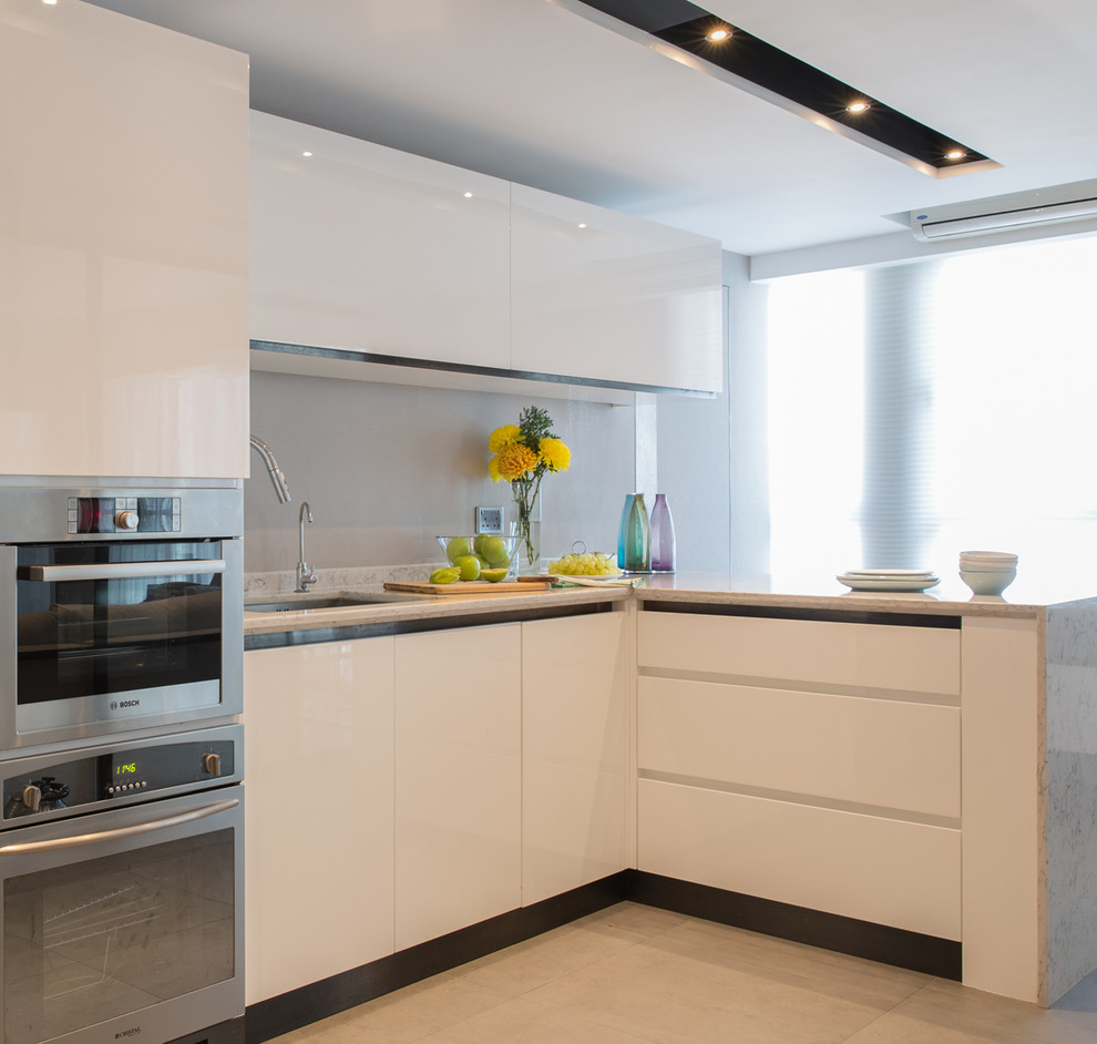 Источник вдохновения для домашнего уюта: угловая, глянцевая кухня-гостиная в современном стиле с врезной мойкой, плоскими фасадами, белыми фасадами, серым фартуком и фартуком из стекла