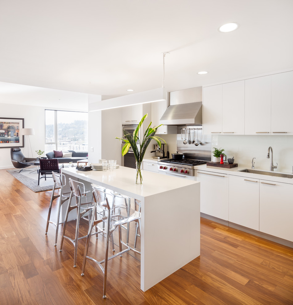 Zweizeilige Moderne Küche mit Unterbauwaschbecken, flächenbündigen Schrankfronten, weißen Schränken, Küchenrückwand in Weiß, Küchengeräten aus Edelstahl, braunem Holzboden und Kücheninsel in Portland