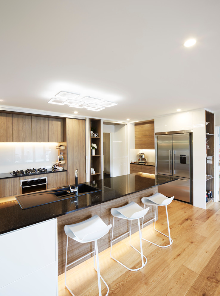 Expansive contemporary u-shaped kitchen/diner in Christchurch with a built-in sink, glass-front cabinets, white cabinets, granite worktops, white splashback, glass sheet splashback, black appliances, plywood flooring and a breakfast bar.