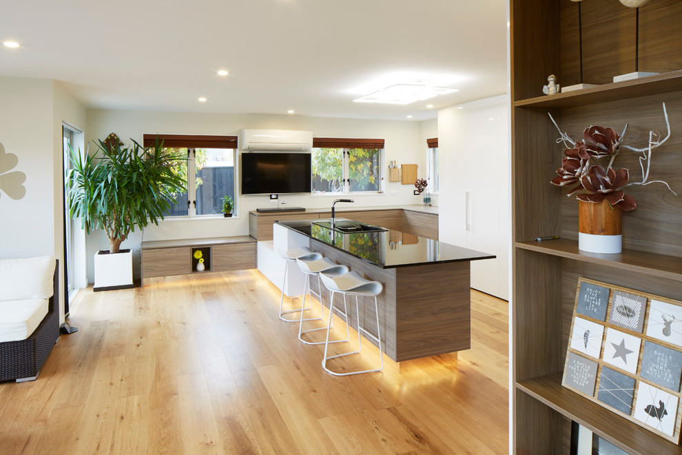 Photo of an expansive contemporary u-shaped kitchen/diner in Christchurch with a built-in sink, glass-front cabinets, white cabinets, granite worktops, white splashback, glass sheet splashback, black appliances, plywood flooring and a breakfast bar.