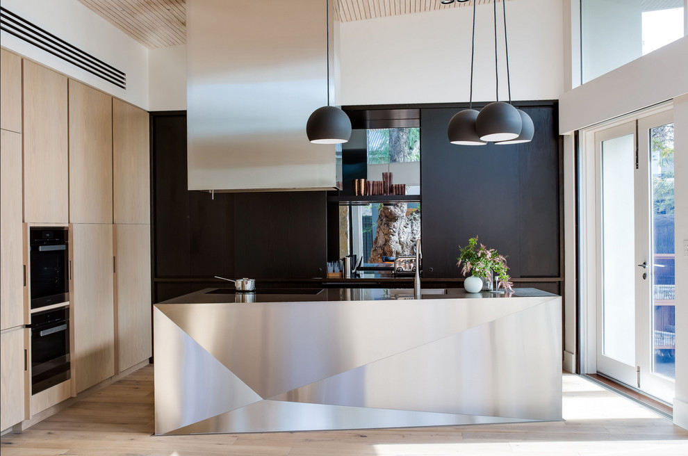 Ejemplo de cocina contemporánea con electrodomésticos negros, suelo de madera clara, una isla, fregadero integrado, armarios con paneles lisos, encimera de acero inoxidable y con blanco y negro