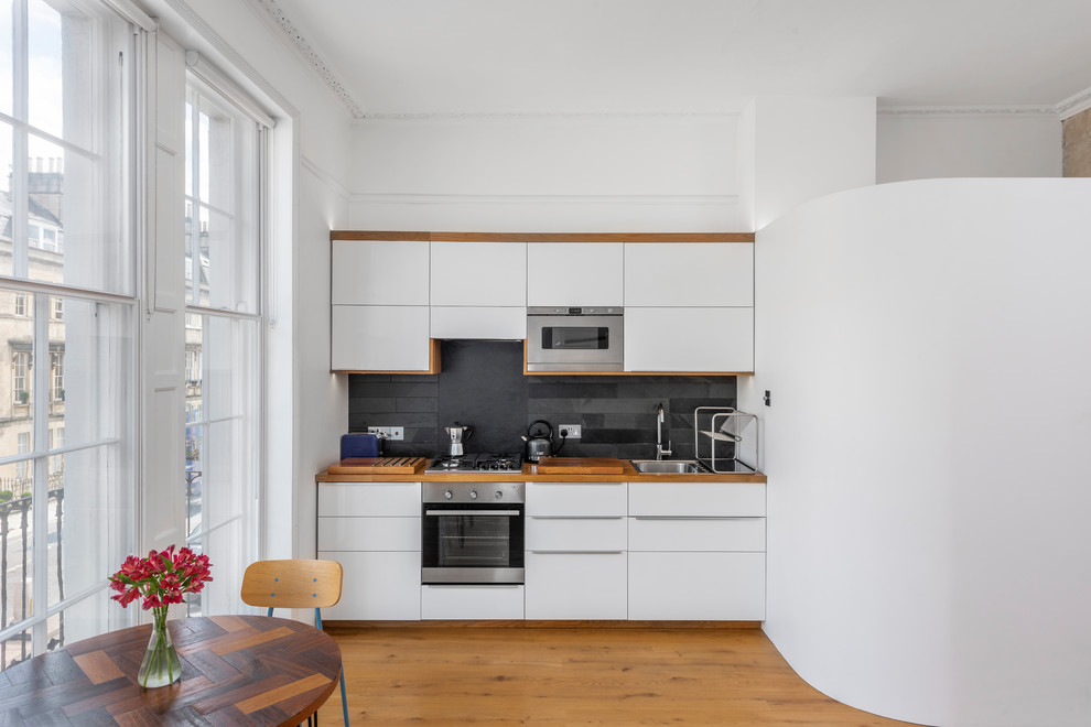 Inspiration for a small contemporary single-wall kitchen/diner in Other with a built-in sink, flat-panel cabinets, white cabinets, wood worktops, slate splashback, black splashback, stainless steel appliances and medium hardwood flooring.