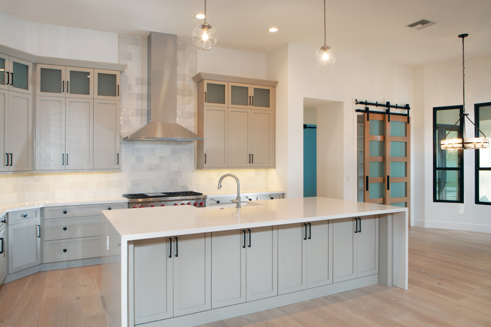 Inspiration for a large classic l-shaped open plan kitchen in Phoenix with a belfast sink, recessed-panel cabinets, grey cabinets, engineered stone countertops, grey splashback, ceramic splashback, stainless steel appliances, light hardwood flooring, an island, beige floors and white worktops.