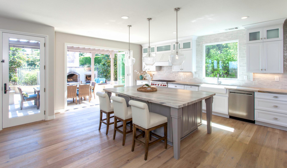 Inspiration for a mid-sized coastal galley laminate floor and brown floor eat-in kitchen remodel in San Diego with white cabinets, quartzite countertops, glass tile backsplash, stainless steel appliances, an island, a farmhouse sink, recessed-panel cabinets, gray backsplash and gray countertops