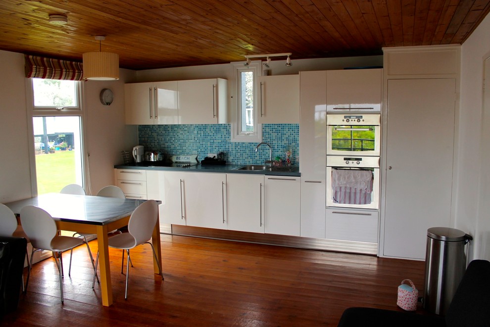 Foto på ett litet maritimt linjärt kök och matrum, med en enkel diskho, släta luckor, vita skåp, laminatbänkskiva, blått stänkskydd, stänkskydd i glaskakel, rostfria vitvaror och mellanmörkt trägolv