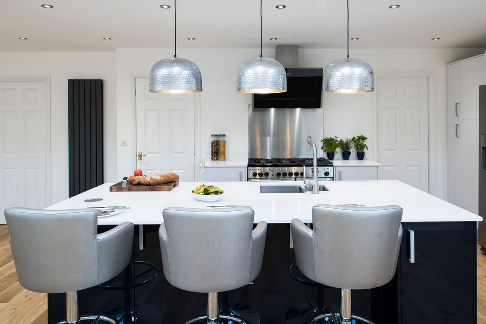 This is an example of a large contemporary l-shaped open plan kitchen in West Midlands with a built-in sink, white cabinets, white splashback, stainless steel appliances, light hardwood flooring, a breakfast bar, beige floors and white worktops.