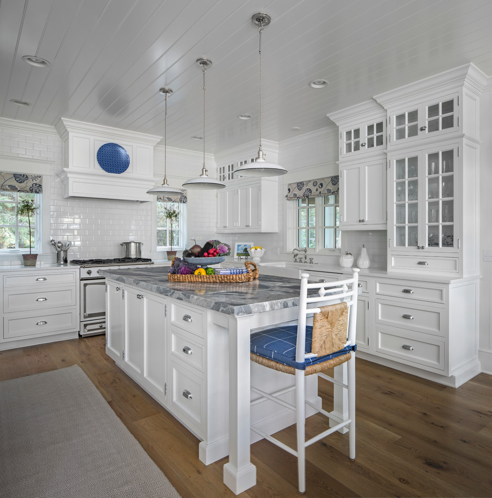 Inspiration for a beach style kitchen in Other with a belfast sink, glass-front cabinets, white cabinets, white splashback, metro tiled splashback, white appliances, light hardwood flooring and an island.
