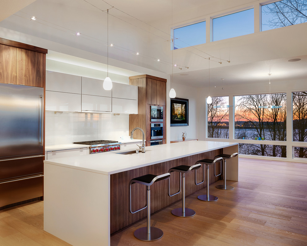 This is an example of a contemporary galley kitchen/diner in Portland with flat-panel cabinets, white cabinets, engineered stone countertops, white splashback, glass sheet splashback, stainless steel appliances, light hardwood flooring, an island and a single-bowl sink.