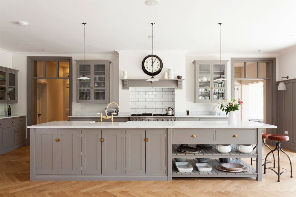 Example of a transitional kitchen design in London with a farmhouse sink, shaker cabinets, gray cabinets, white backsplash, subway tile backsplash, stainless steel appliances and an island