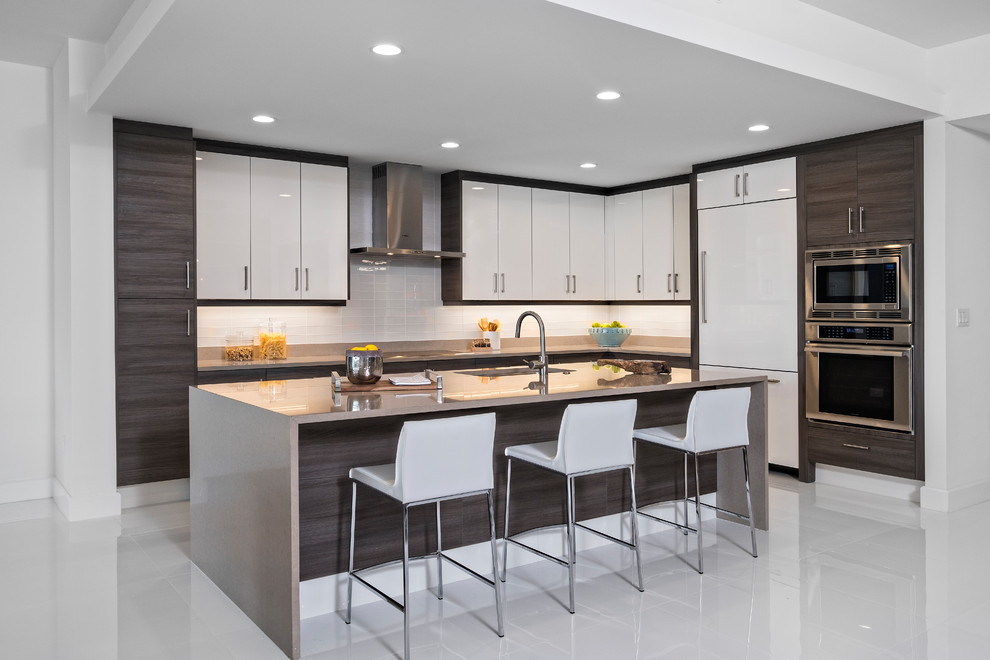 Contemporary l-shaped kitchen in Miami with a submerged sink, flat-panel cabinets, white cabinets, grey splashback, glass tiled splashback, integrated appliances, an island, white floors and grey worktops.