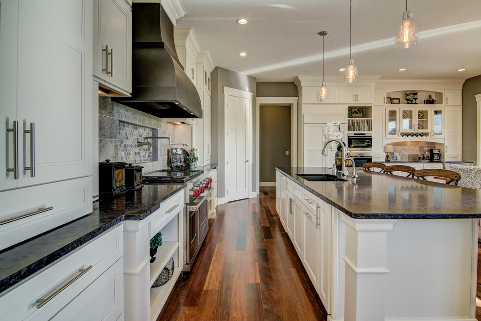 Klassisk inredning av ett stort kök, med en undermonterad diskho, luckor med infälld panel, vita skåp, granitbänkskiva, grått stänkskydd, stänkskydd i stenkakel, rostfria vitvaror, mörkt trägolv och en köksö